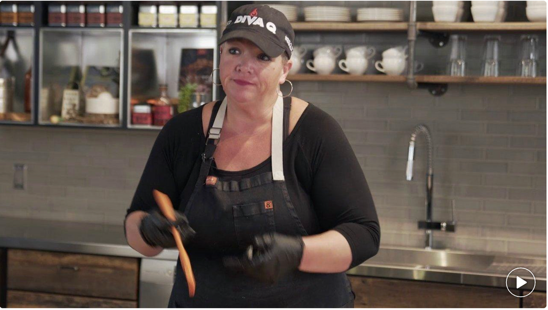 Person in a kitchen wearing a black apron and gloves, holding a wooden spoon.