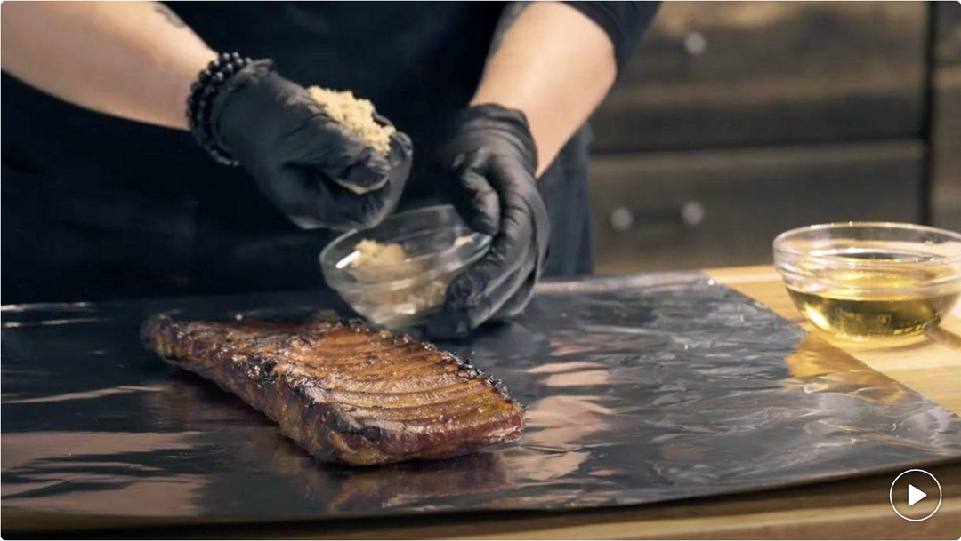 Person seasoning a steak with a bowl of spices on a dark surface.