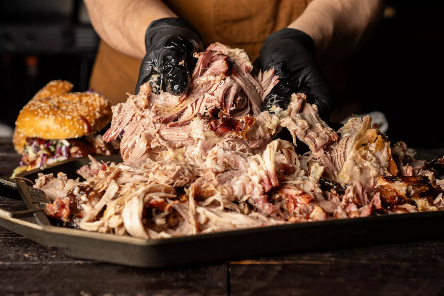 Person holding pulled pork over a tray with a sandwich in the background